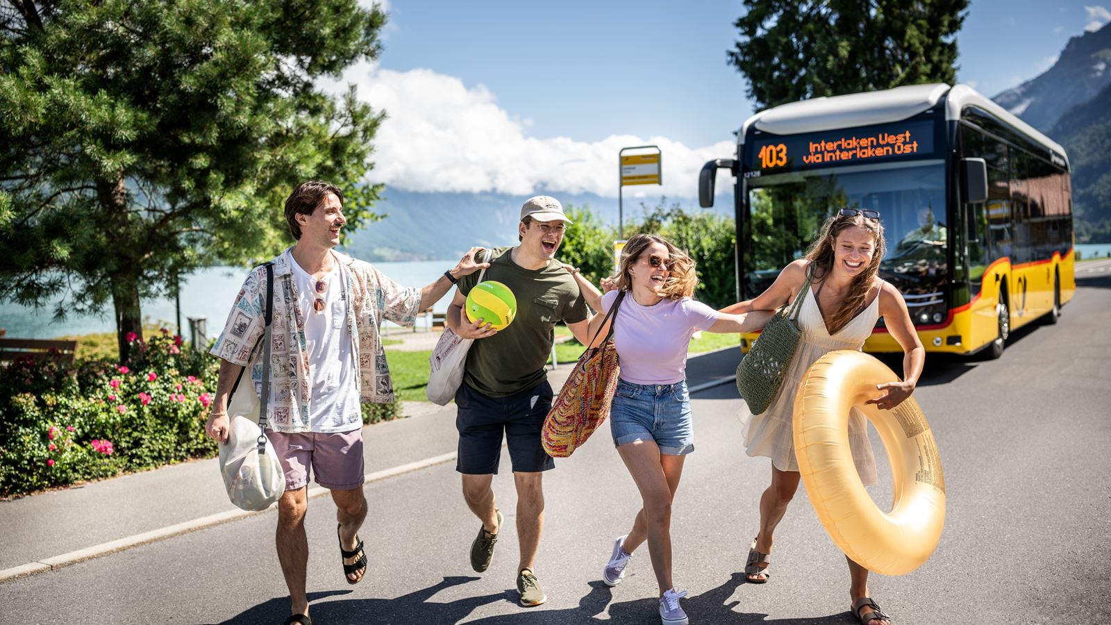 A group of young people with bathing utensils crossing a road by Lake Thun in front of a Postbus.