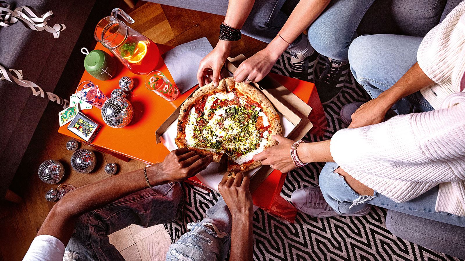 Sicht von oben auf Hände von drei Personen, die ein Stück Pizza auf einem Salontisch greifen. Auf dem Tisch stehen neben der Pizza noch Getränke und klein Diskokugeln.