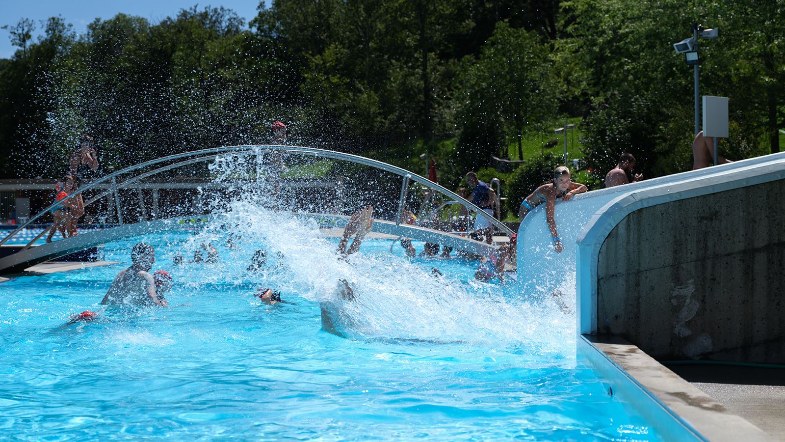 Hauptbecken des Schwimmbads Tesserete mit einem Kind, das von der Rutsche ins Wasser eintaucht.