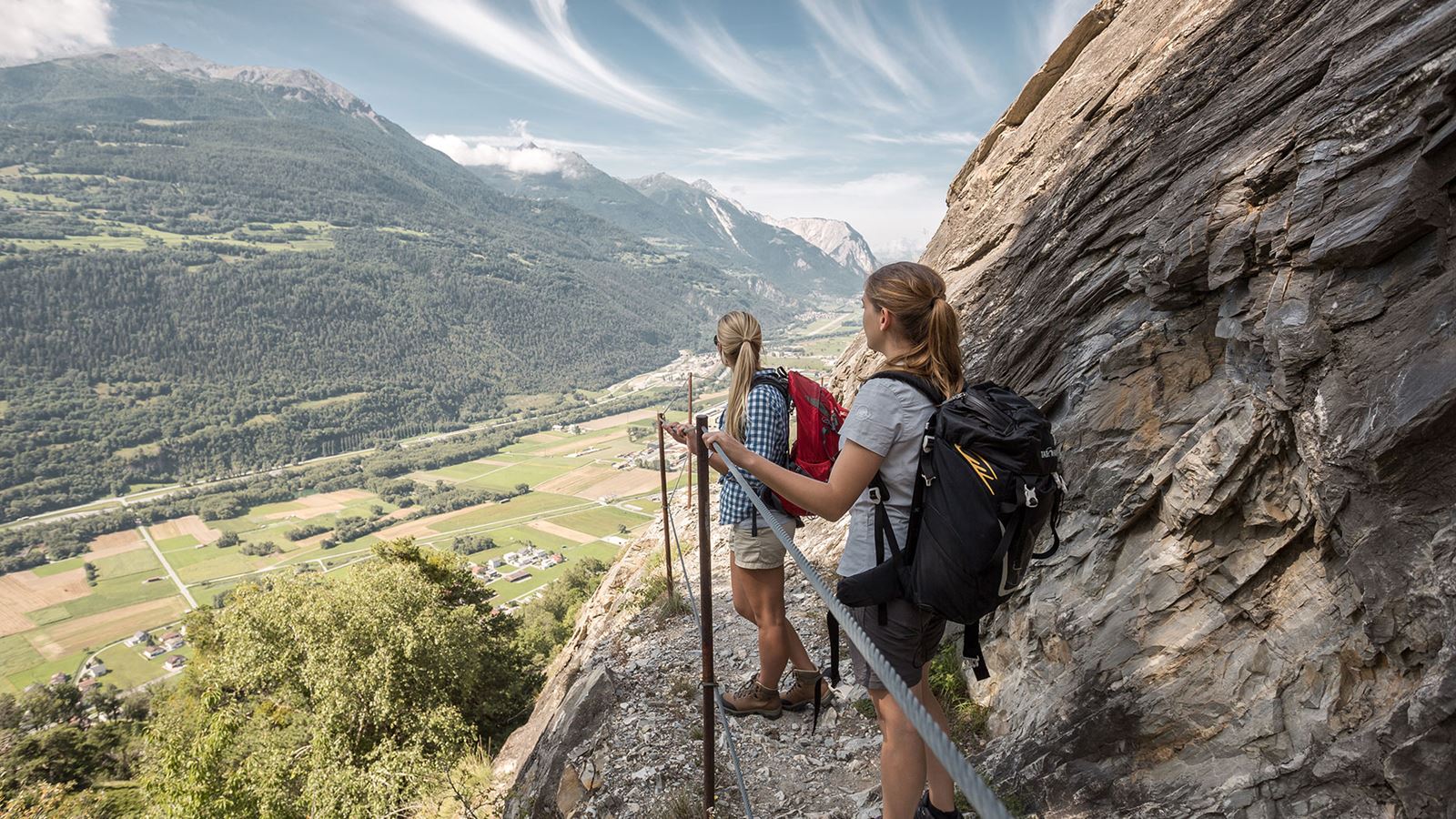Zwei Wandernde mit Rucksäcken gehen auf einem schmalen, gesicherten Bergpfad entlang einer Felswand und schauen über ein Tal mit Feldern und Bergen im Hintergrund.