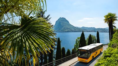 Postauto fährt entlang des Luganersees in Richtung St.Moritz mit dem Monte Bre im Hintergrund. 