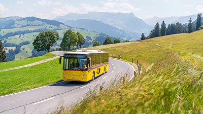 Postauto fährt auf Strasse in ländlicher Umgebung. Im Hintergrund die Hügellandschaft vom Entlebuch mit Blick auf die Schrattenfluh.