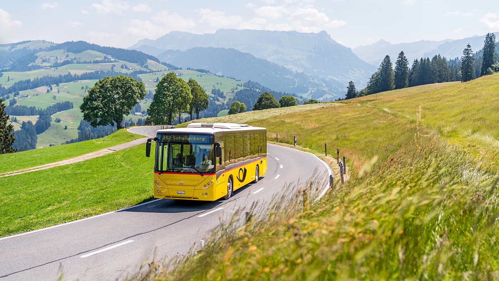 Postauto fährt auf Strasse in ländlicher Umgebung. Im Hintergrund die Hügellandschaft vom Entlebuch mit Blick auf die Schrattenfluh.