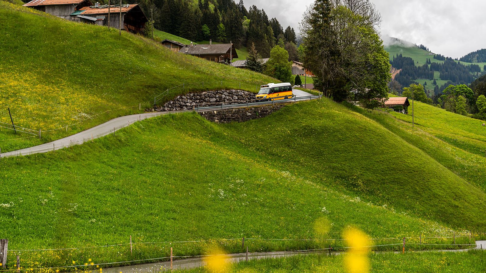 Un petit CarPostal descend l’étroite route. En arrière-plan, on aperçoit des maisons traditionnelles et des prairies verdoyantes.