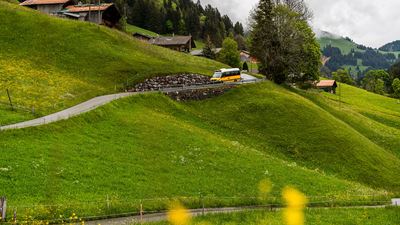 Un petit CarPostal descend l’étroite route. En arrière-plan, on aperçoit des maisons traditionnelles et des prairies verdoyantes.
