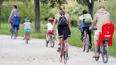 Persone adulte e bambini pedalano tra gli alberi verdi che circondano l’aeroporto di Zurigo.