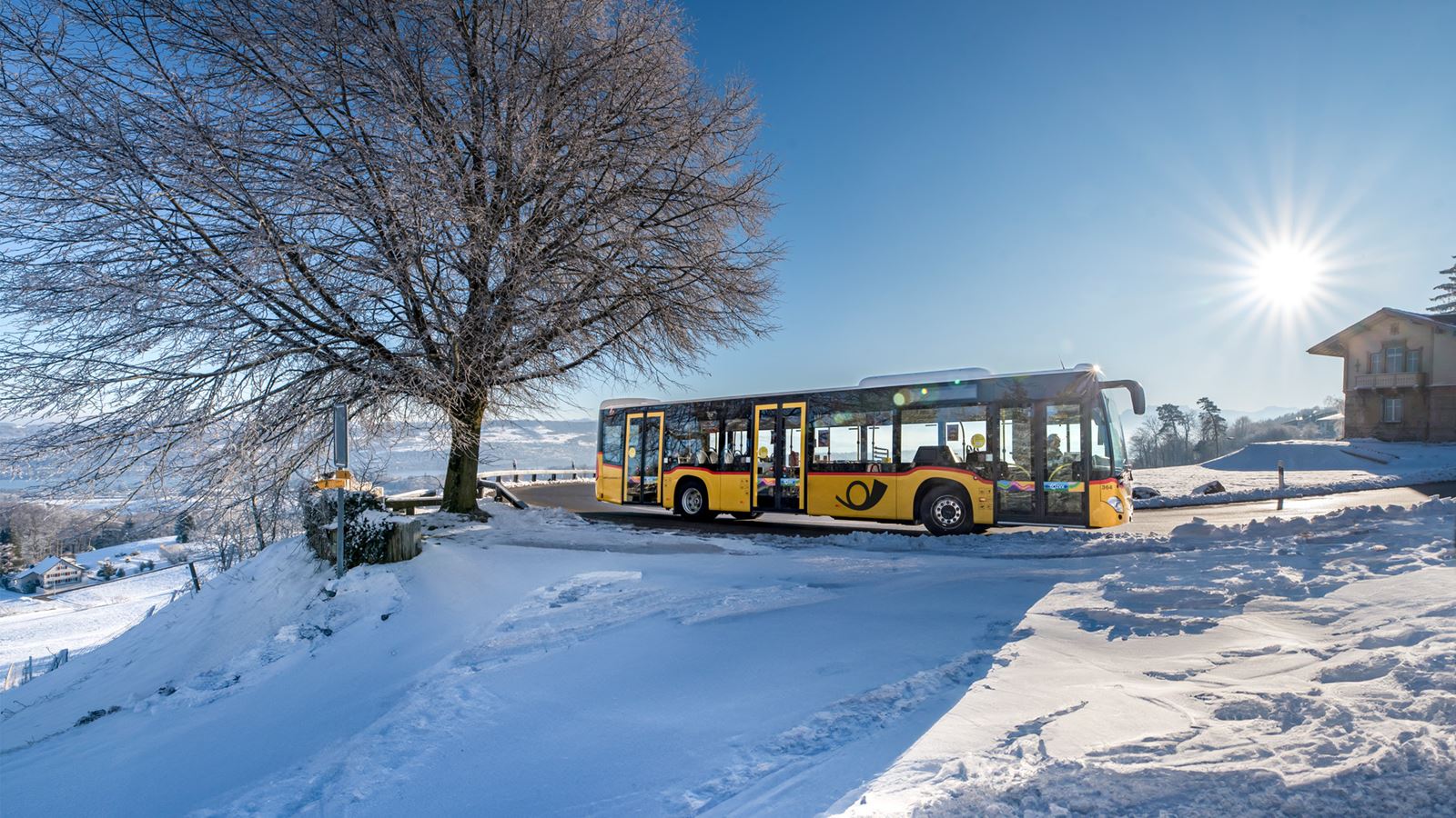 Un car postal jaune sillonne le paysage enneigé du col de l’Albis sous un soleil radieux.