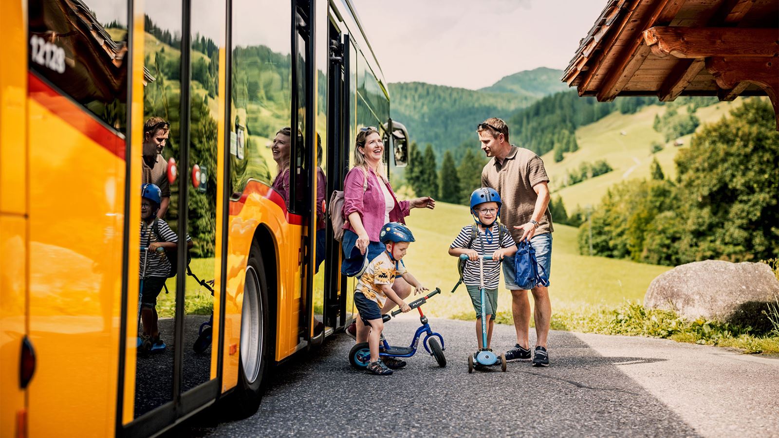 Two adults and two children wearing helmets get off a PostBus on a country road in a green, hilly landscape.