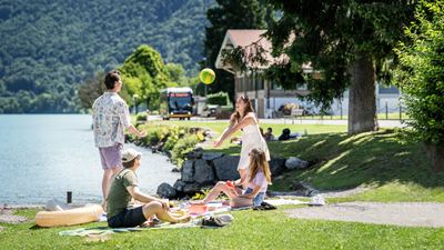 Quatre personnes font un pique-nique au bord du lac et jouent avec une balle; en arrière-plan, il y a des arbres, une maison et un car postal.