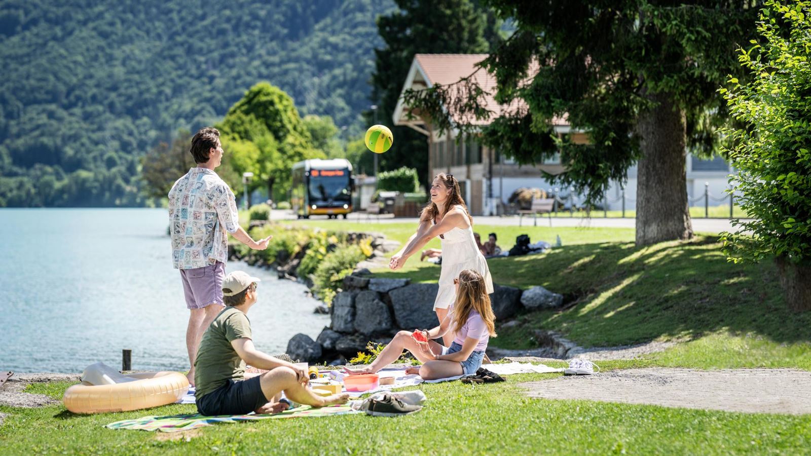 Four people have a picnic on the lake shore and play with a ball; in the background are trees, a house and a Postbus.