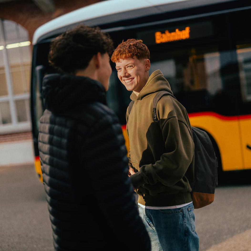 Un jeune homme souriant se tient devant un car postal portant l’inscription «Gare». Au premier plan figure un autre entretien avec une personne portant un pull à capuche. L’arrière-plan montre la façade d’un bâtiment. L’atmosphère est conviviale et décontractée.