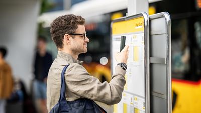 Ein Mann steht an einer PostAuto-Haltestelle und fotografiert mit seinem Smartphone den Fahrplan. Im Hintergrund ist ein PostAuto zu sehen. Die Atmosphäre ist urban und modern.