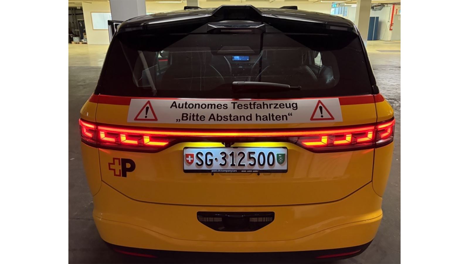 A yellow AmiGo vehicle from behind. Above the number plate, there is a poster labelled “Autonomous test vehicle, please keep your distance!”.
