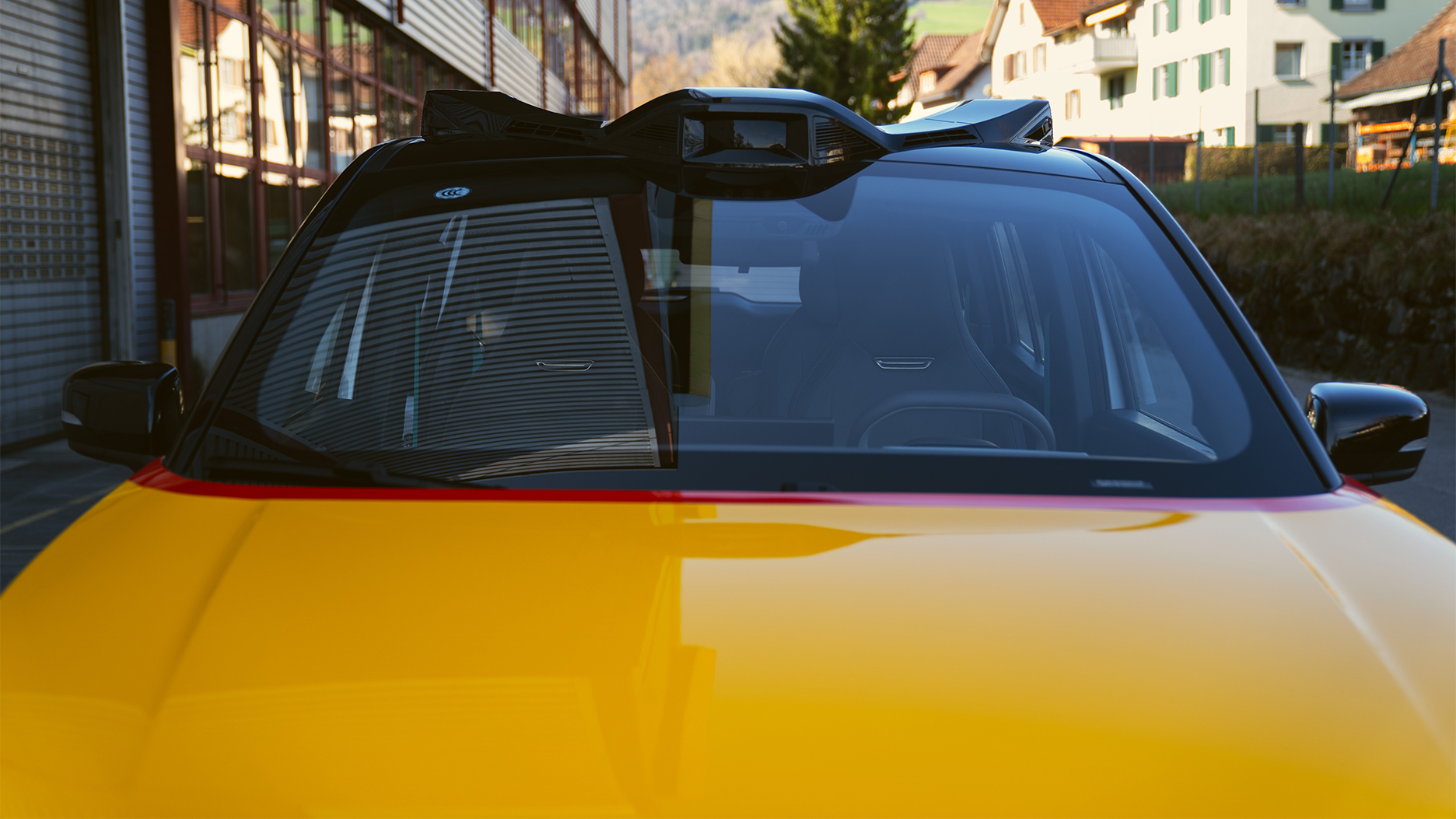 Close-up of a yellow AmiGo vehicle with visible sensors on the roof; residential buildings and landscape in the background.
