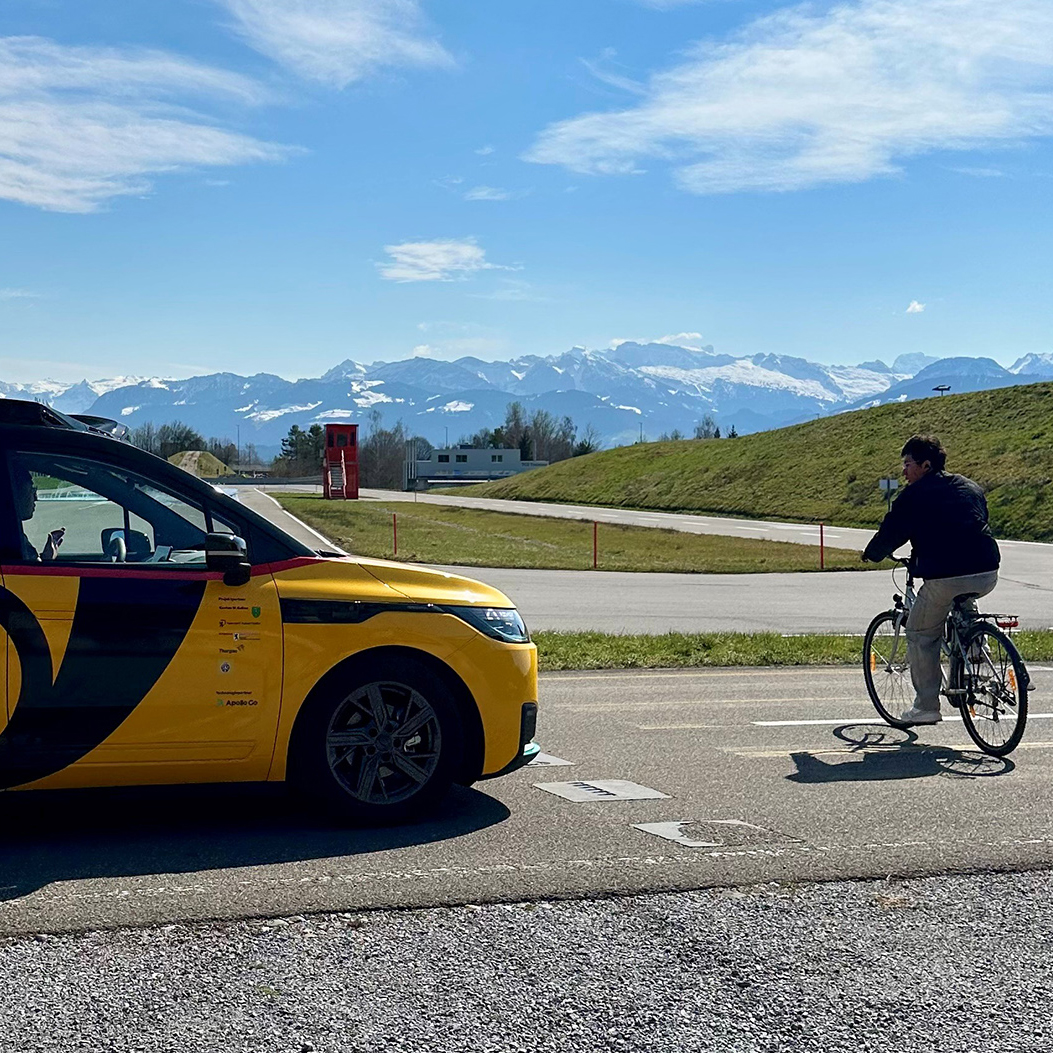 Yellow AmiGo vehicle stops on test track while a person crosses the road on a bike; mountains can be seen in the background.