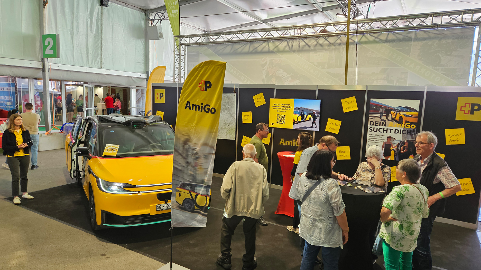 The AmiGo yellow vehicle at the trade fair stand stands with doors open in a hall, with information walls and visitors in the background.