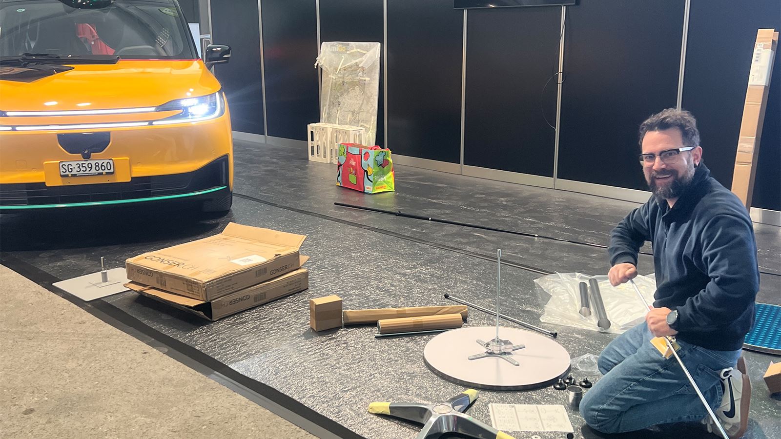 Allestimento di uno stand fieristico: il veicolo AmiGo giallo è pronto, una persona monta il materiale espositivo sulla superficie di vendita.