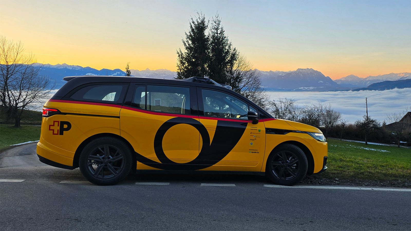 Véhicule jaune autonome AmiGo stationné sur le bord de la route devant un panorama alpin avec une mer de brouillard en arrière-plan en Suisse.