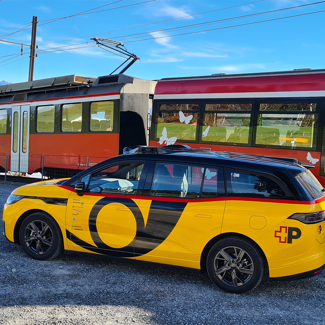AmiGo Fahrzeug steht neben einem Zug an einer Bahnstrecke im ländlichen Raum.