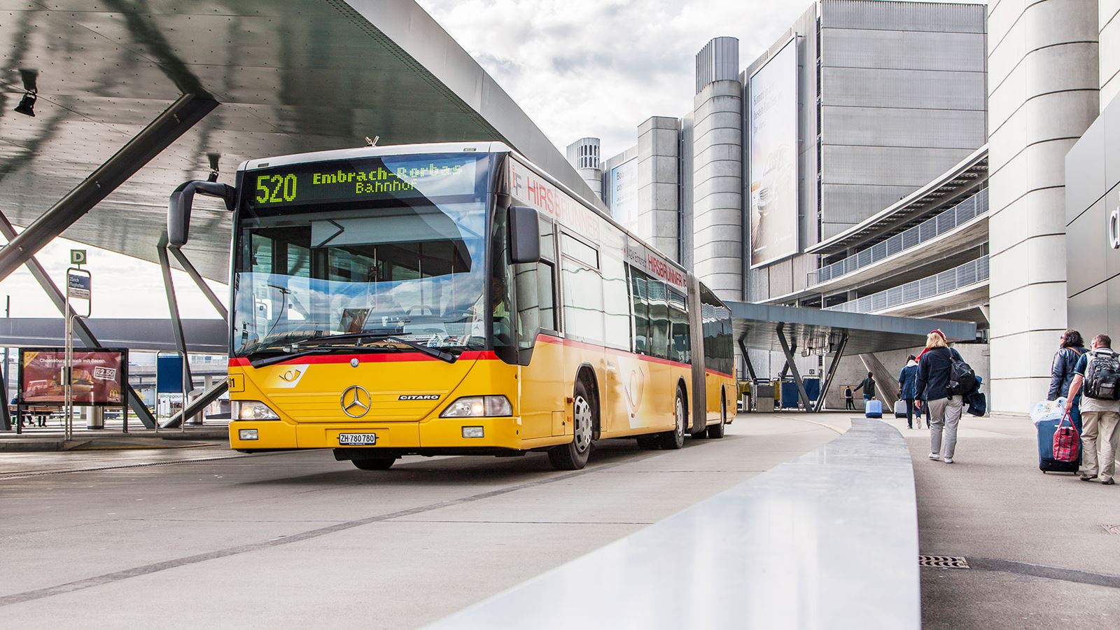 Un car postal s’arrête à une gare dans un environnement urbain.