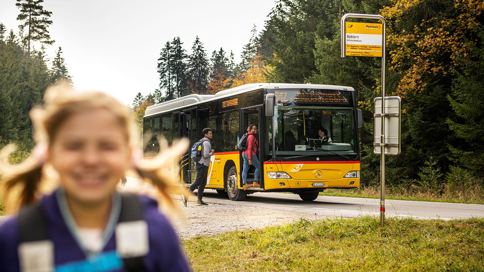 There is a girl’s head in the foreground, and two passengers boarding a Postbus at a stop in the background. 