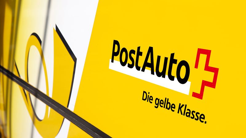 Gros plan sur le flanc d’un car postal où l’on voit le nom de l’entreprise, le cor postal et le slogan «La classe jaune». 