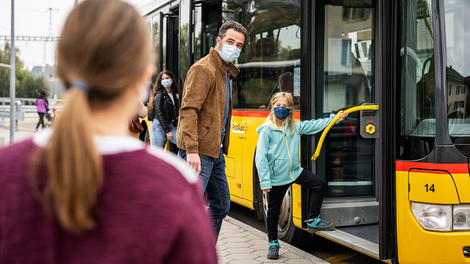 An adult man and a child boarding a Postbus. Both are wearing masks on their faces. 